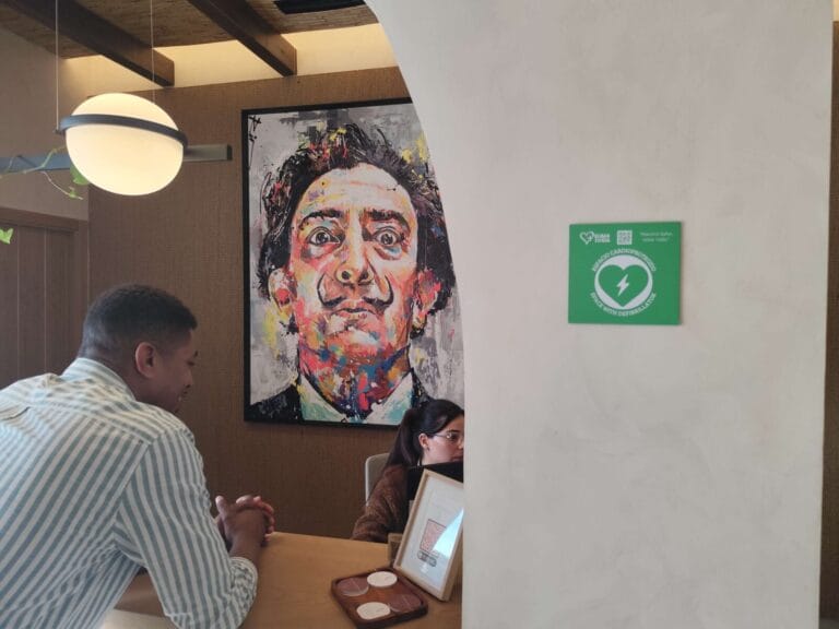 Placa verde de espacio cardioprotegido con desfibrilador visible en la pared de la recepción del hotel, con un cuadro de Salvador Dalí al fondo.